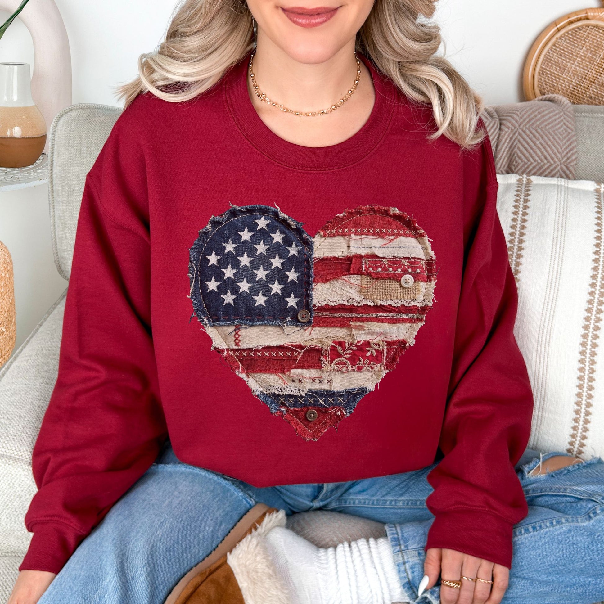Model wearing a Gildan 18000 Sweatshirt in muted denim blue (washed denim) with a patriotic heart design made of red, white, and blue American flag patterns in a faux patchwork and stitched fabric style.