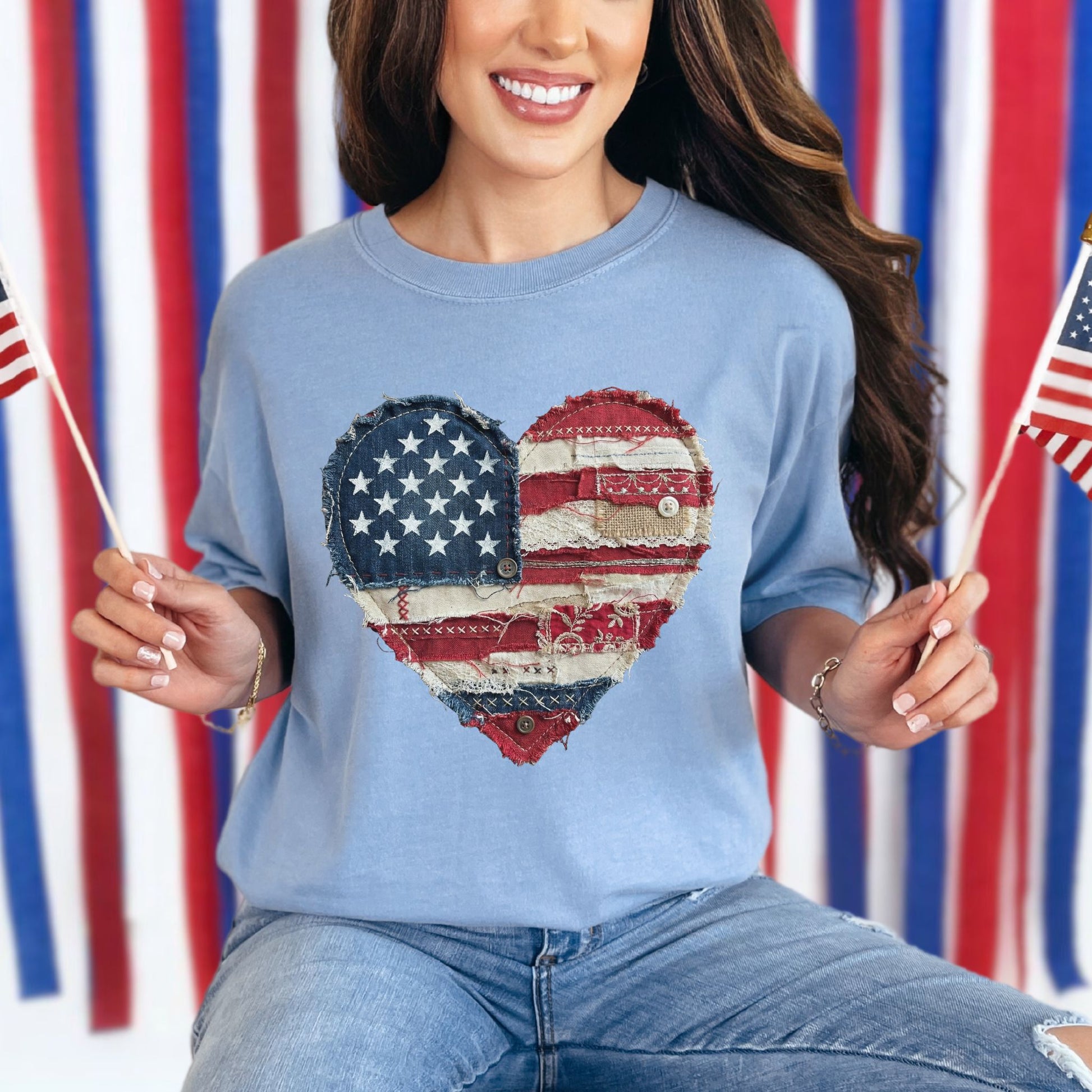 Model wearing a Comfort Colors 1717 t-shirt in muted denim blue (washed denim) with a patriotic heart design made of red, white, and blue American flag patterns in a faux patchwork and stitched fabric style.