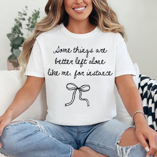 Model wearing a Comfort Colors 1717 t-shirt in bright white (white) color featuring a graphic of a hand written font saying "Some things are better left alone like me, for instance”. Underneath the phrase is a bow.