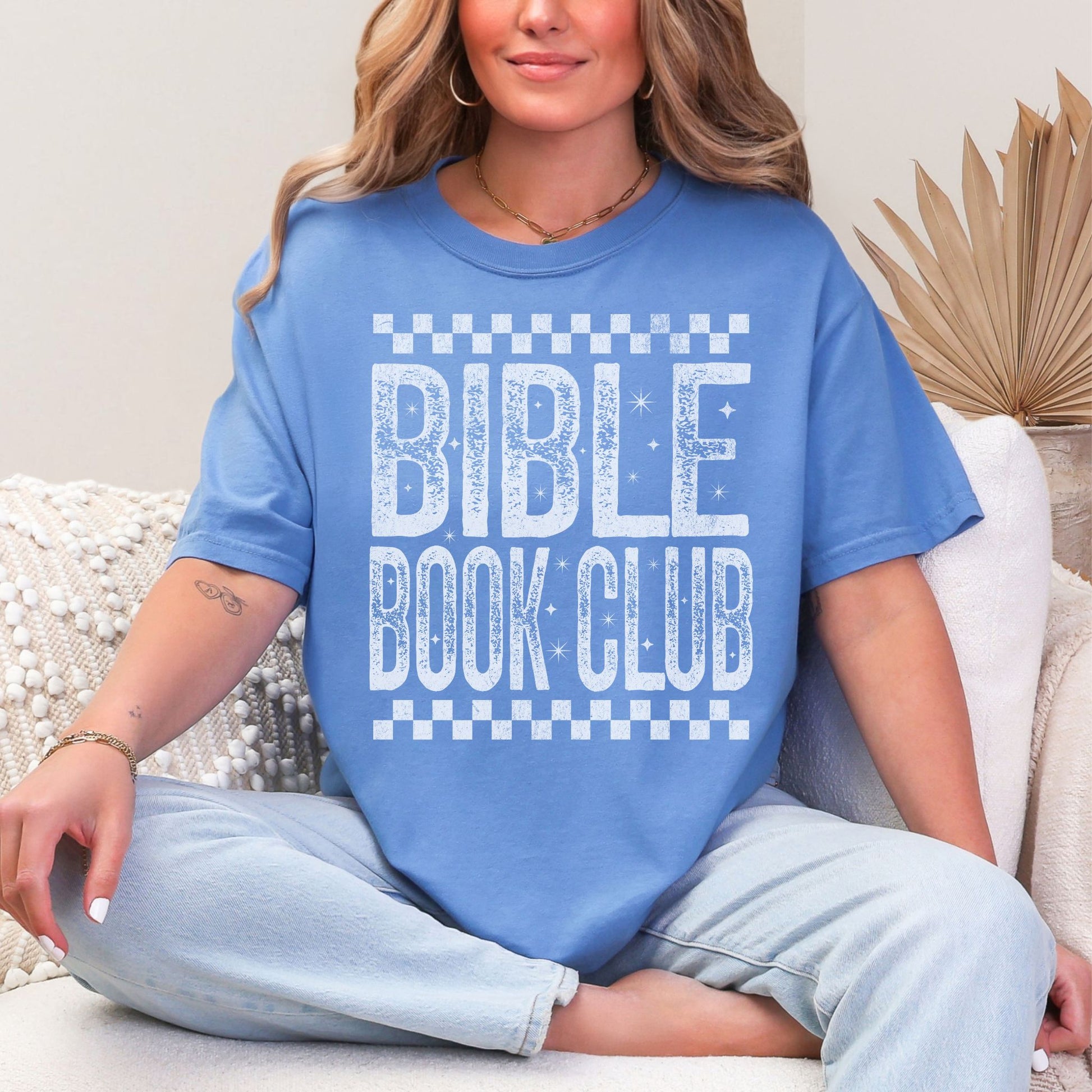 Model wearing a Comfort Colors 1717 T-Shirt in muted blue-green (mystic blue) featuring a distressed large word graphic that says, "Bible Book Club" Racer checkered stripes are on the top and bottom of the print.
