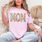 Model is wearing a Comfort Colors t-shirt in soft light pink (blossom) featuring the word “mom” with “blessed” in script across it in a floral faux embroidery style design.