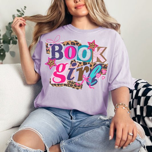 Model wearing a Comfort Colors 1717 t-shirt in soft purple (orchid) featuring a colorful ‘Book Girl’ collage graphic with leopard print accents, stars, bows, and hearts.