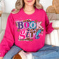 Model wearing a Gildan 18000 on bright pink (heliconia) featuring a colorful ‘Book Girl’ collage graphic with leopard print accents, stars, bows, and hearts.