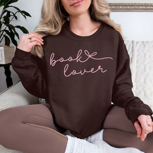 Model wearing a Gildan 18000 sweatshirt in a deep dark brown (dark chocolate) color featuring a graphic design of a cursive script font that says "book lover" with a bow after book.