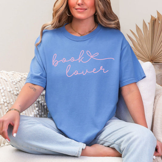 Model wearing a Comfort Colors 1717 t-shirt in a muted blue-green (mystic blue) color featuring a graphic design of a cursive script font that says "book lover" with a bow after book.