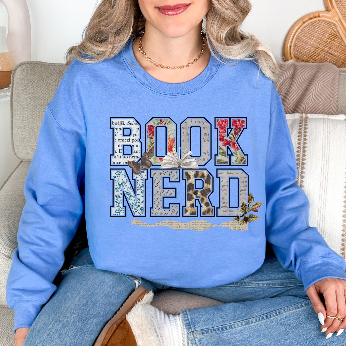 Model wearing a Gildan 18000 soft sky blue (carolina blue) Sweatshirt featuring the words “BOOK NERD” in large block letters filled with collage-style patterns including vintage book pages, florals, and textures, with an open book and small botanical accents incorporated into the design.