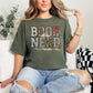 Model wearing a Comfort Colors 1717 Muted green (sage) T-shirt featuring the words “BOOK NERD” in large block letters filled with collage-style patterns including vintage book pages, florals, and textures, with an open book and small botanical accents incorporated into the design.