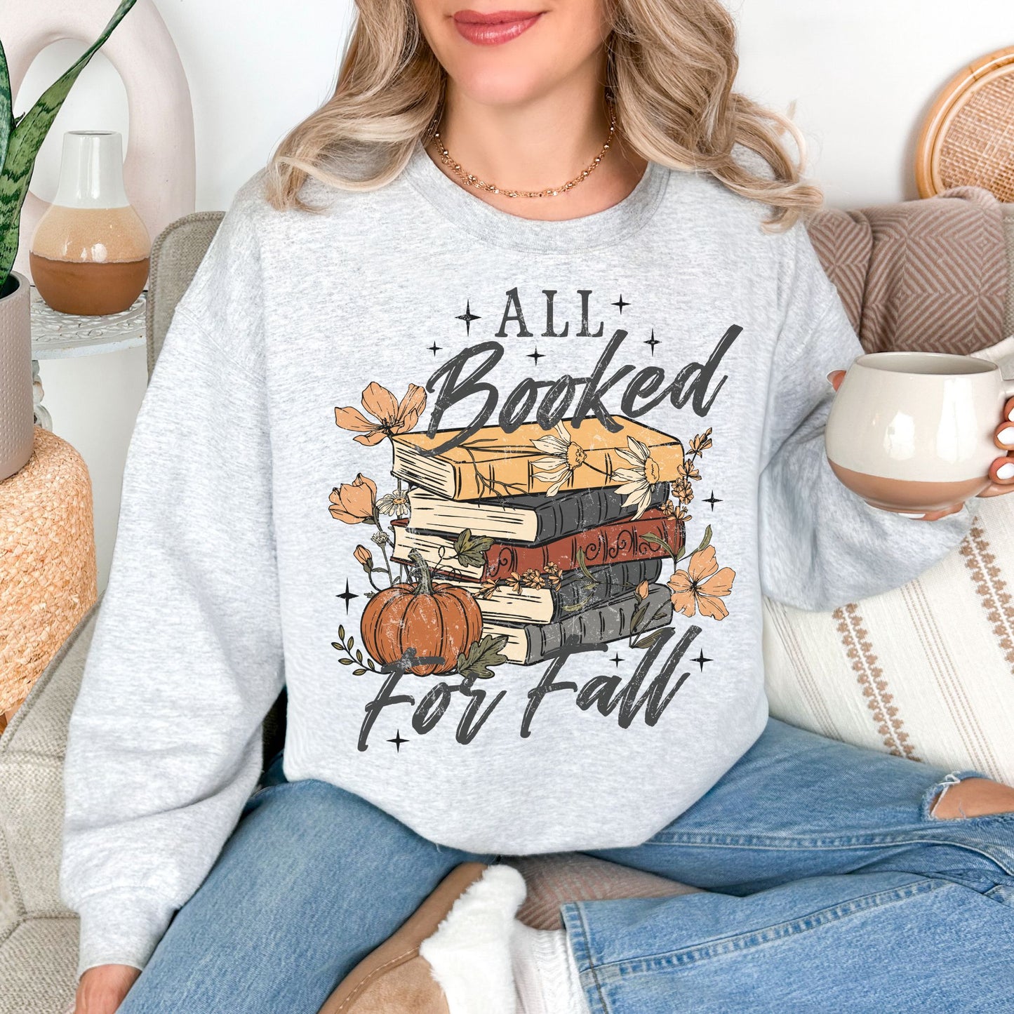 Model wearing a Gildan 18000 sweatshirt in very light gray (ash) featuring an “All Booked for Fall” design with stacked books, pumpkins, autumn flowers, leaves, and stars.