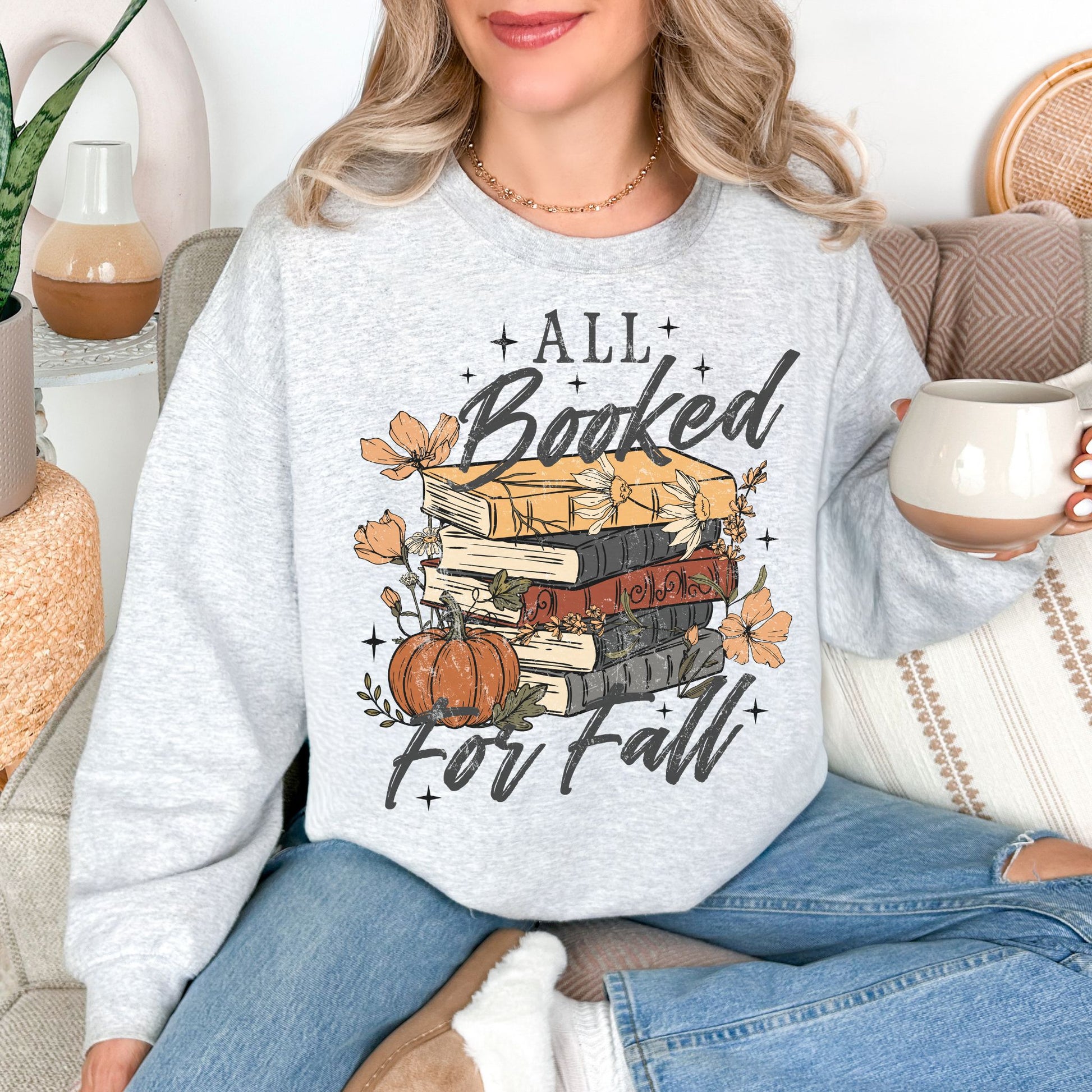 Model wearing a Gildan 18000 sweatshirt in very light gray (ash) featuring an “All Booked for Fall” design with stacked books, pumpkins, autumn flowers, leaves, and stars.