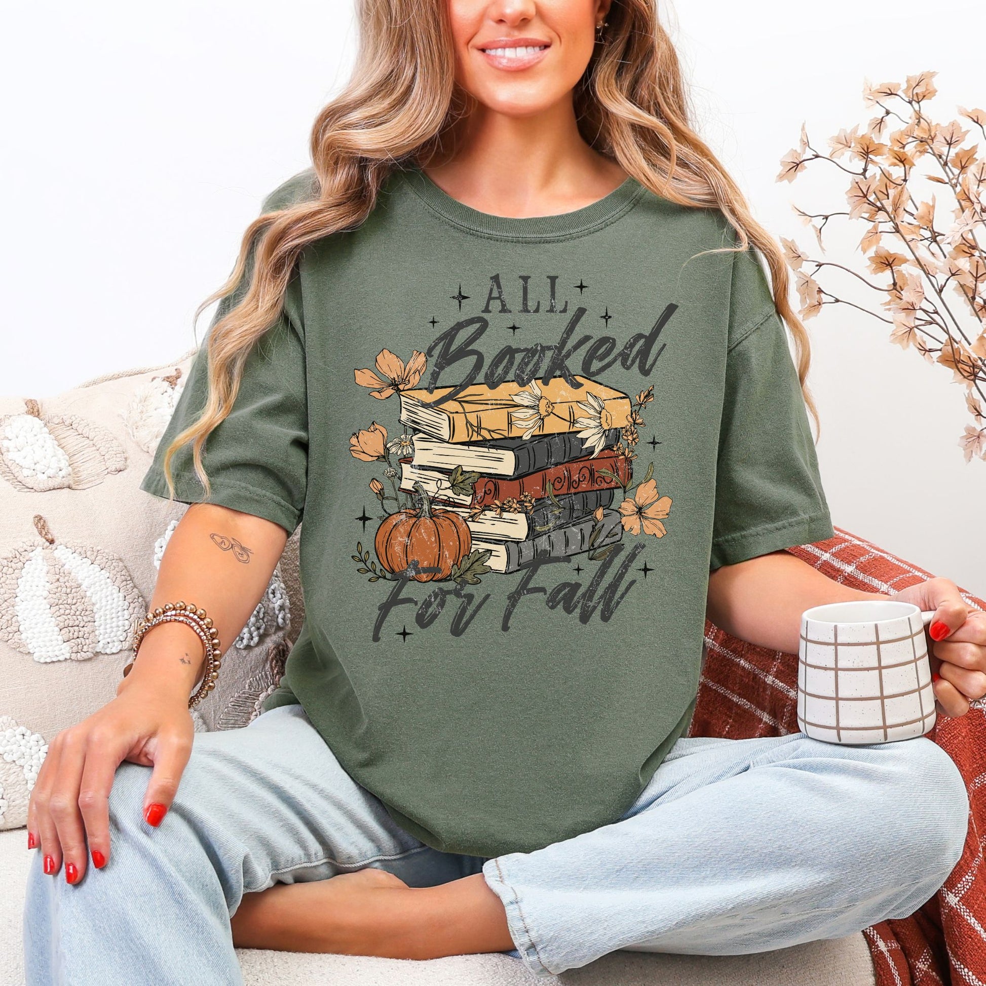 Model wearing a Comfort Colors 1717 t-shirt in dark muted green (moss) featuring an “All Booked for Fall” design with stacked books, pumpkins, autumn flowers, leaves, and stars.