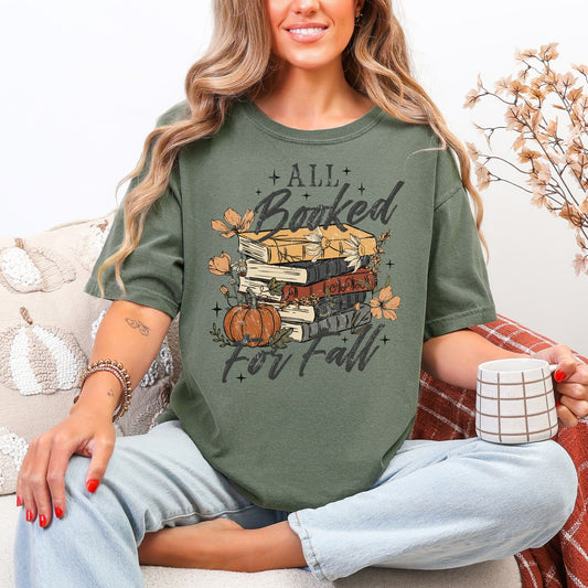 Model wearing a Comfort Colors 1717 t-shirt in dark muted green (moss) featuring an “All Booked for Fall” design with stacked books, pumpkins, autumn flowers, leaves, and stars.