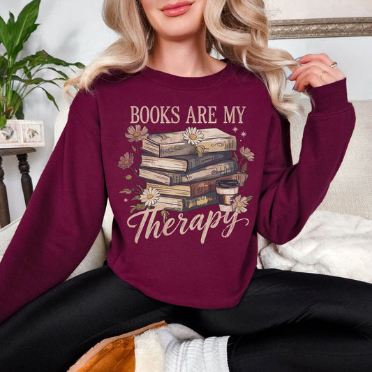 Model wearing a Gildan 18000 Sweatshirt in a muted deep burgundy red (maroon) color featuring a stacked book graphic with a to-go coffee and daisies. The phrase around the graphic is "Books are my therapy".