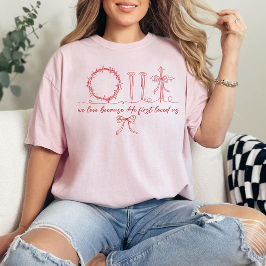 Model wearing a Comfort Colors soft pink (blossom) T-Shirt featuring a red hand-drawn / whimsical devotional sketch of a ring of thorns, nails, and a cross. The wording "we love because He first loved us" underneath in a beautiful cursive font. Then a doodle sketch bow is underneath to bring the graphic together. 