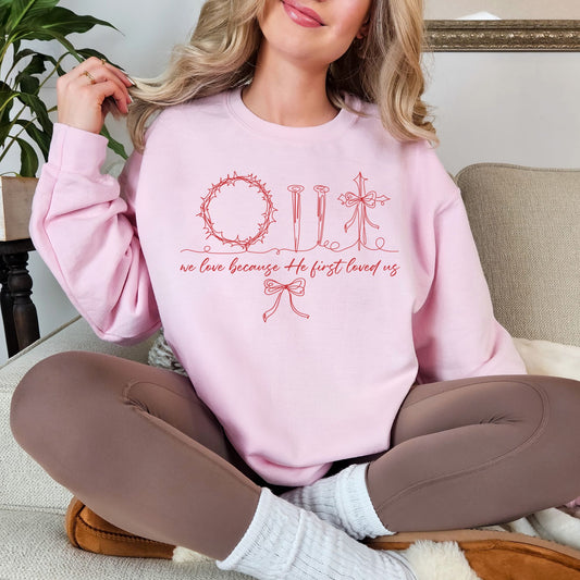 Model wearing a Gildan 18000 very soft pale pink (light pink) sweatshirt featuring a red hand-drawn / whimsical devotional sketch of a ring of thorns, nails, and a cross. The wording "we love because He first loved us" underneath in a beautiful cursive font. Then a doodle sketch bow is underneath to bring the graphic together. 