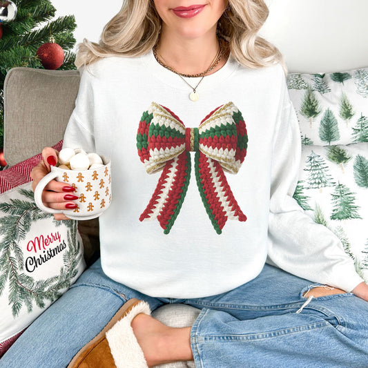 Model wearing a Gildan 18000 long-sleeve sweatshirt in white (White) featuring a large Christmas bow with red, green, and cream faux crochet stitches, gold-colored trim, and two long ribbon tails.