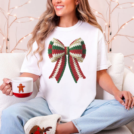 Model wearing a Comfort Colors 1717 short-sleeve t-shirt in white (White) featuring a large Christmas bow with red, green, and cream faux crochet stitches, gold-colored trim, and two long ribbon tails.