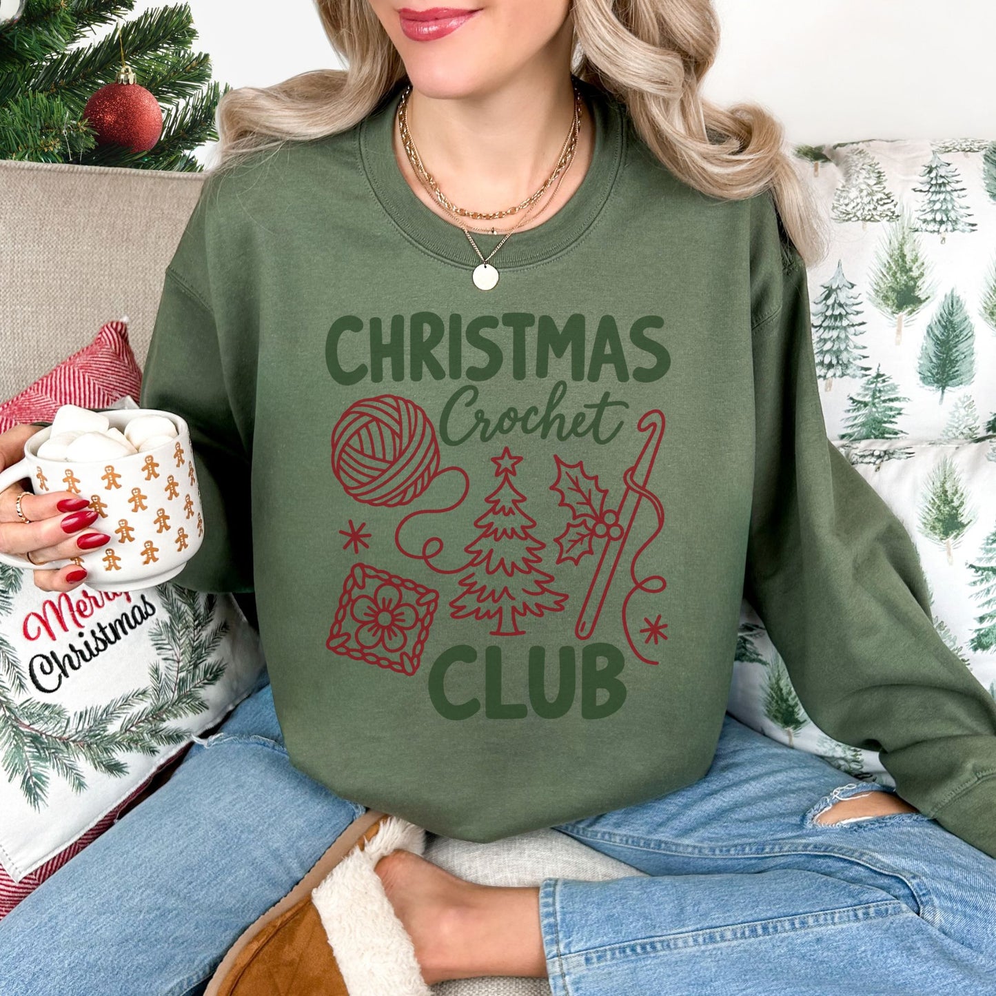 Model wearing a Gildan 18000 long sleeve sweatshirt in muted olive green (military green) featuring the words “Christmas Crochet Club” with illustrations of a yarn ball, crochet hook, Christmas tree, holly leaves, stars, and a floral crochet square.