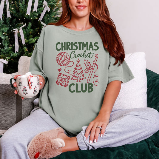 Model wearing a Comfort Colors® 1717 short-sleeve t-shirt in light gray-green (Bay) featuring the words “Christmas Crochet Club” with illustrations of a yarn ball, crochet hook, Christmas tree, holly leaves, stars, and a floral crochet square.