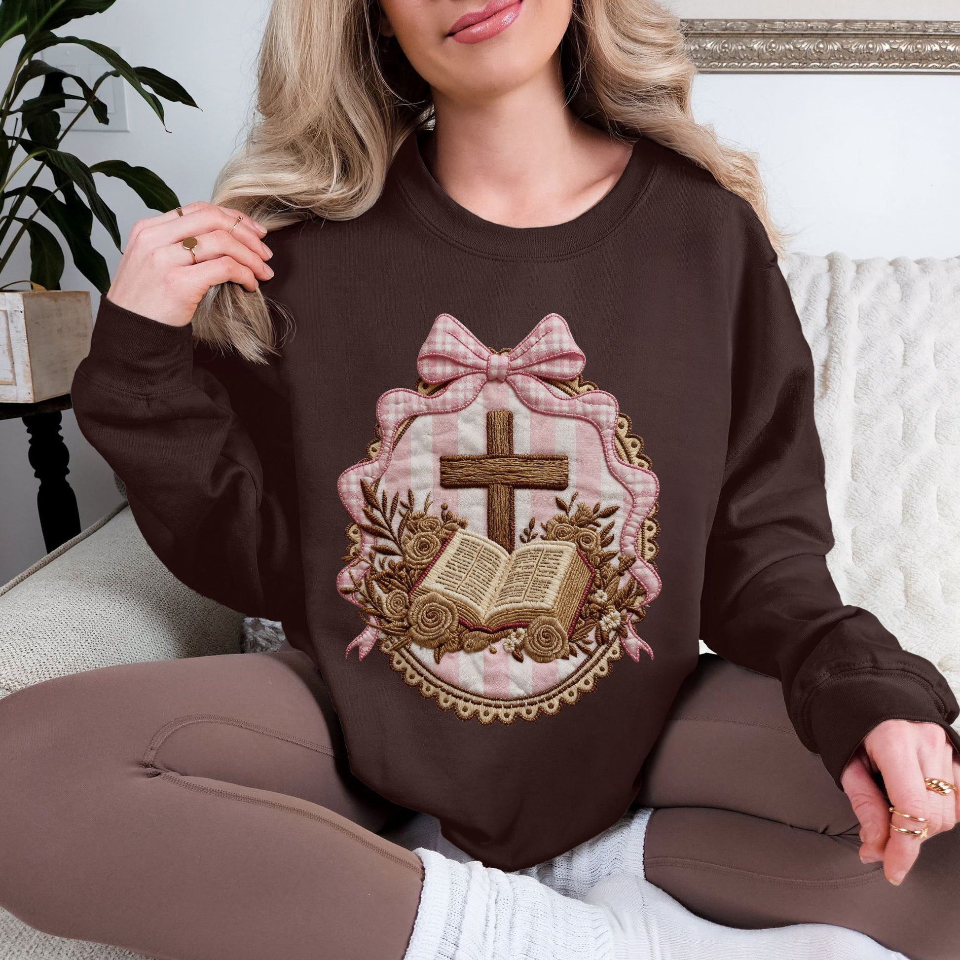 Model wearing a Gildan 18000 sweatshirt in deep dark brown (dark chocolate) featuring a faux embroidery-style illustration of a wooden cross above an open Bible, surrounded by floral accents and framed with pink striped fabric and a pink bow. The design is printed artwork made to resemble embroidery and stitched fabric but is not real stitching or textured appliqué.