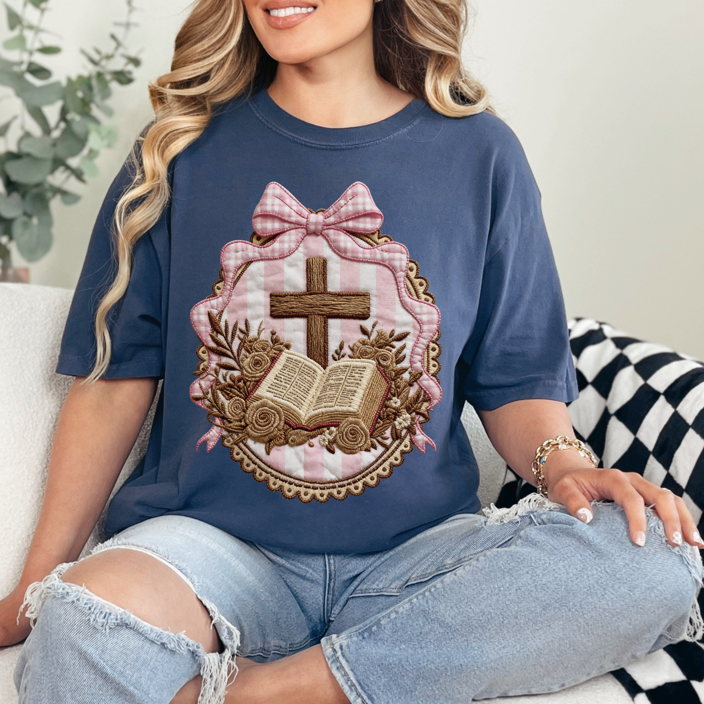 Model wearing a Comfort Colors 1717 T-shirt in deep navy blue (navy) featuring a faux embroidery-style illustration of a wooden cross above an open Bible, surrounded by floral accents and framed with pink striped fabric and a pink bow. The design is printed artwork made to resemble embroidery and stitched fabric but is not real stitching or textured appliqué.