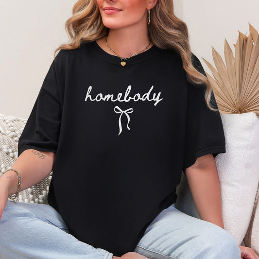 Model wearing a Comfort Colors T- shirt in black featuring a handwriting script graphic of the word "Homebody". Below the script is a tiny bow.