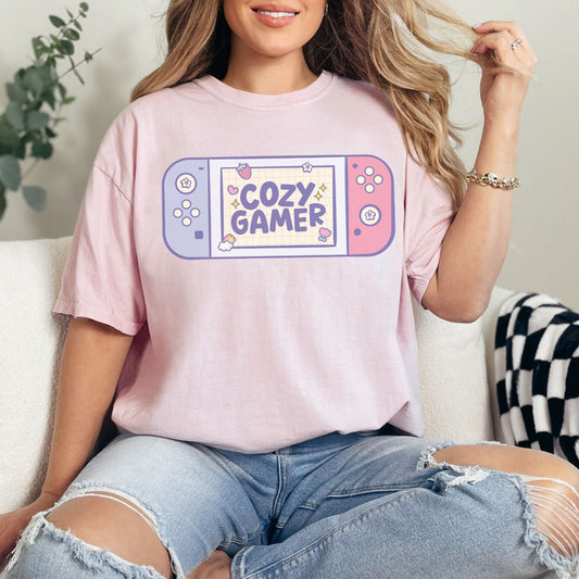 Model wearing a Comfort Colors 1717 t-shirt in a soft pink (blossom) color featuring a pastel illustrated handheld gamer console. Inside the illustration are the words "Cozy Gamer".