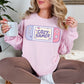 Model wearing a Gildan 18000 Sweatshirt in soft pale pink (light pink) color featuring a pastel illustrated handheld gamer console. Inside the illustration are the words "Cozy Gamer"