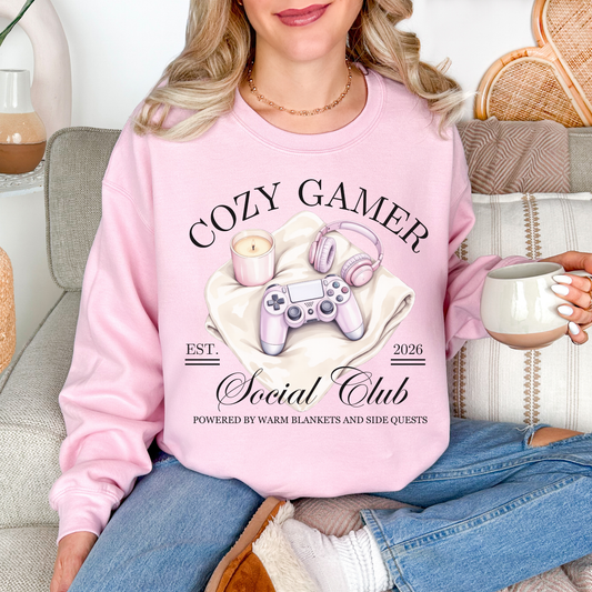 Model wearing a Gildan 18000 Sweatshirt in a soft pale pink (light pink) color featuring a cozy gamer graphic with a controller, candle and a headset. It writing above the graphic says, "Cozy Gamer". Below, it says "Social Club powered by warm blankets and side quests" 