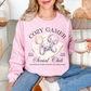 Model wearing a Gildan 18000 Sweatshirt in a soft pale pink (light pink) color featuring a cozy gamer graphic with a controller, candle and a headset. It writing above the graphic says, "Cozy Gamer". Below, it says "Social Club powered by warm blankets and side quests" 