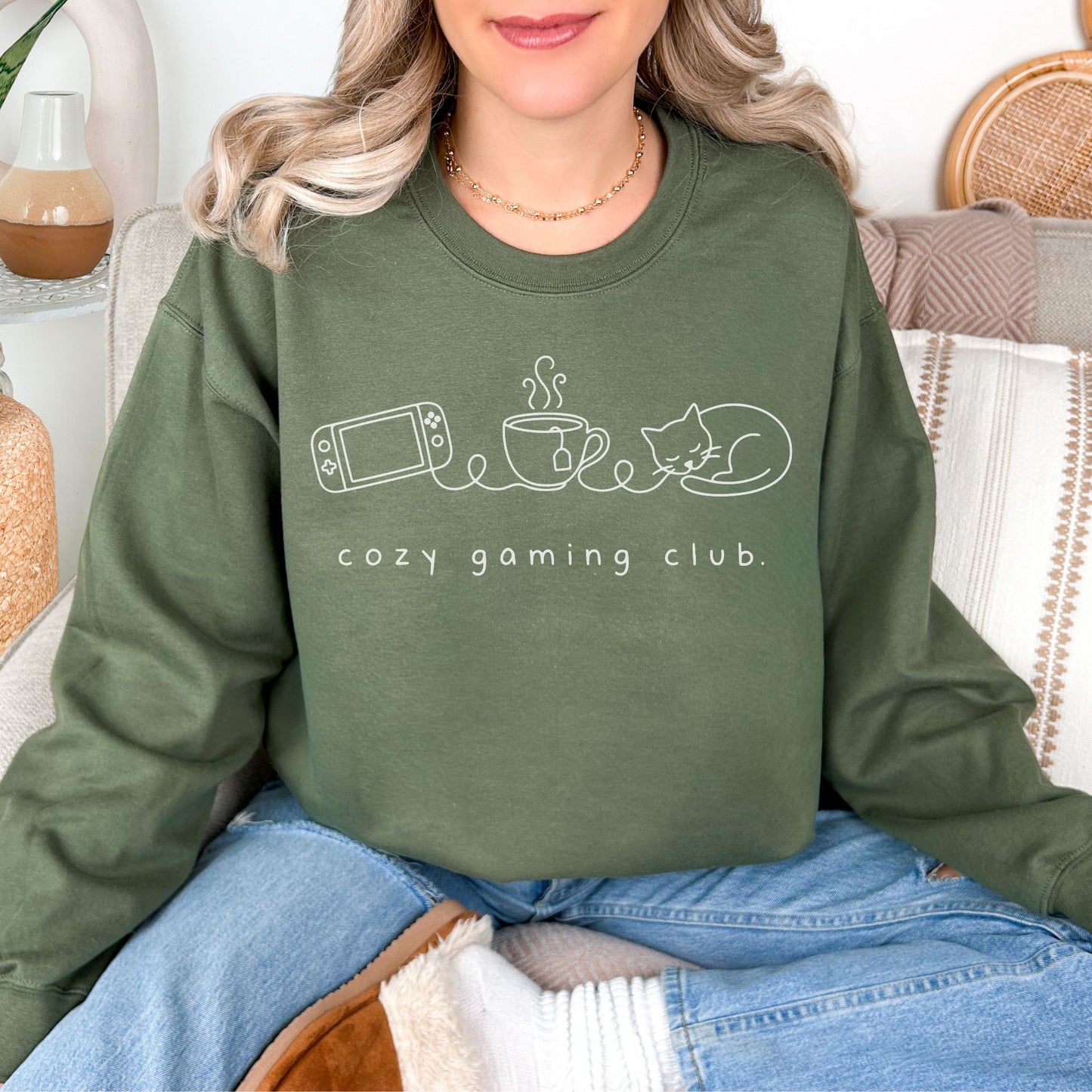Model wearing a Gildan 18000 sweatshirt in muted olive green (military green) featuring a line art graphic of a gaming console, a cup of tea, and a sleeping cat. Underneath are the words "cozy gaming club."