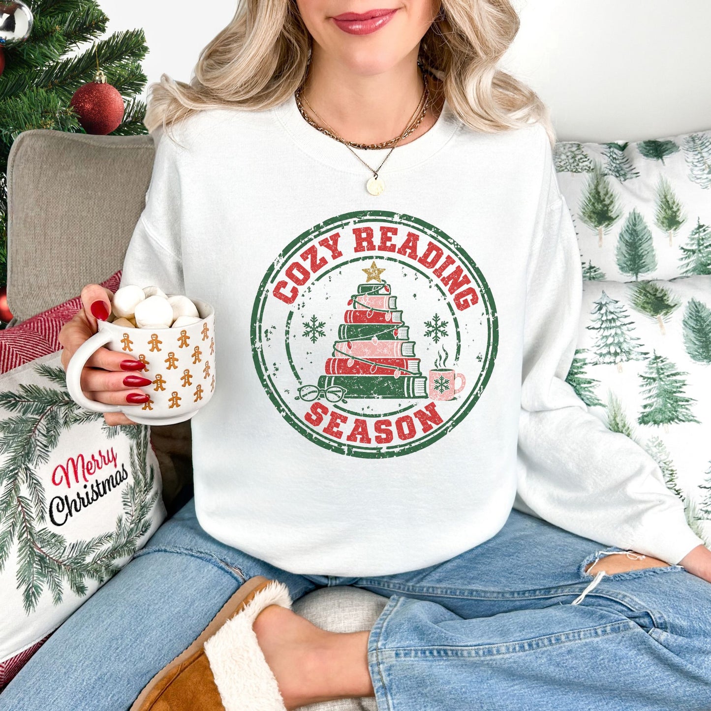 Model wearing a Gildan 18000 sweatshirt in white (White) featuring a distressed circular Christmas graphic with the words “Cozy Reading Season” in red lettering, a green border, a stack of red and green books arranged as a Christmas tree, a gold star, green snowflakes, reading glasses, and a pink steaming mug.