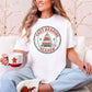 Model wearing a Comfort Colors® 1717 short-sleeve t-shirt in white (White) featuring a distressed circular Christmas graphic with the words “Cozy Reading Season” in red lettering, a green border, a stack of red and green books arranged as a Christmas tree, a gold star, green snowflakes, reading glasses, and a pink steaming mug.