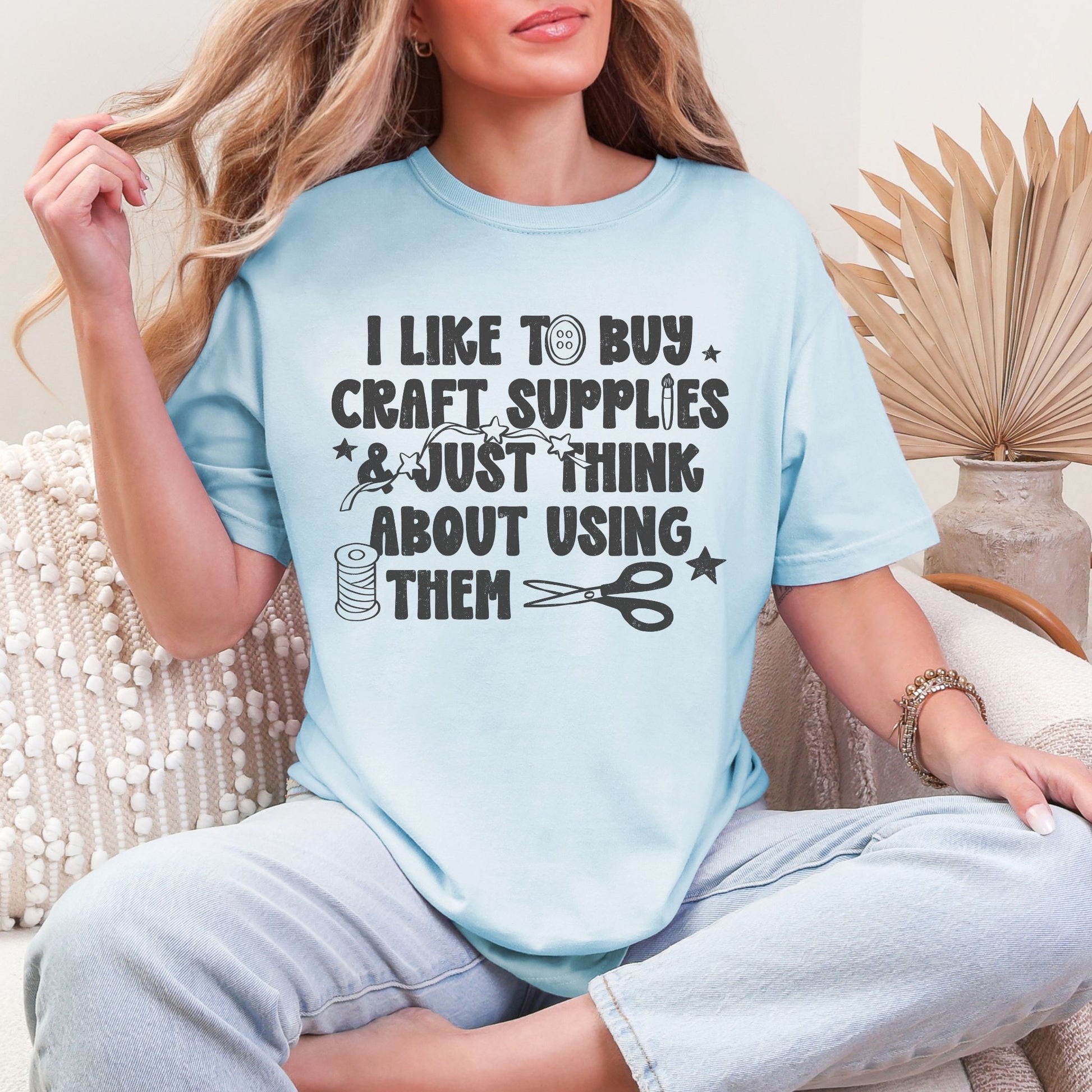 Model is wearing a Comfort Colors 1717 shirt in soft medium blue (chambray) featuring a funny craft-themed design that reads "I Like to Buy Craft Supplies & Just Think About Using Them" with sewing-inspired graphics including thread, scissors, stars, and buttons.