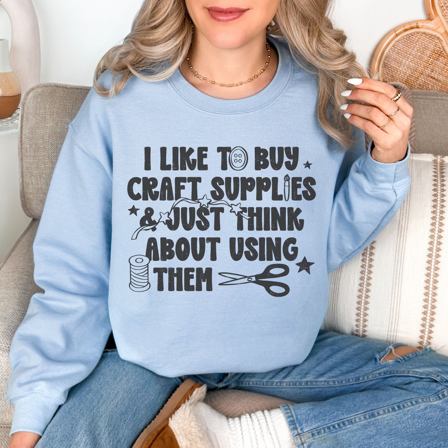 Model is wearing a Comfort Colors 1717 sweatshirt in pale blue (light blue) featuring a funny craft-themed design that reads "I Like to Buy Craft Supplies & Just Think About Using Them" with sewing-inspired graphics including thread, scissors, stars, and buttons.