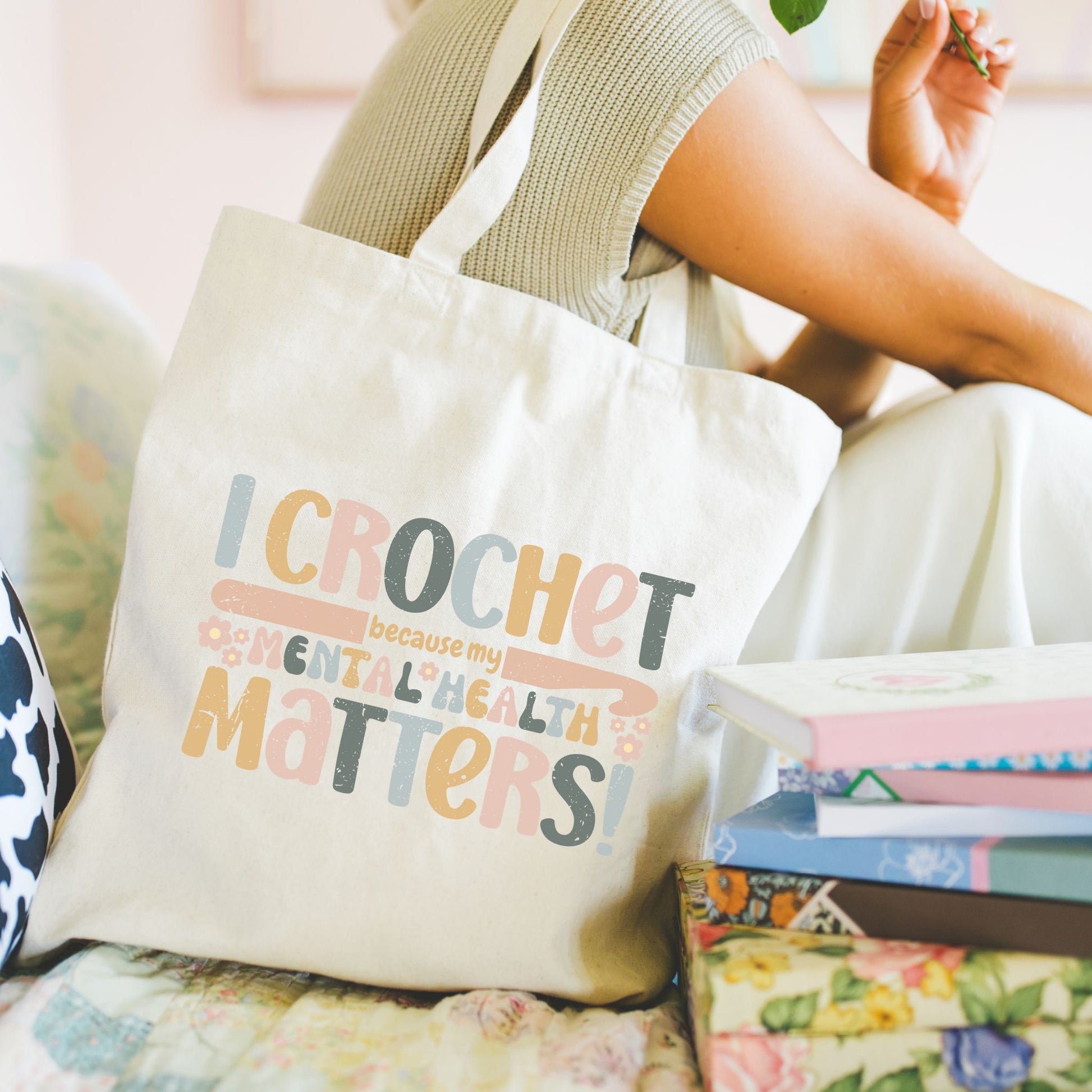 Model sitting with a Natural cotton canvas tote bag featuring the phrase “I crochet because my mental health matters!” in soft pastel lettering with small floral accents in peach, yellow, sage green, and muted blue tones.