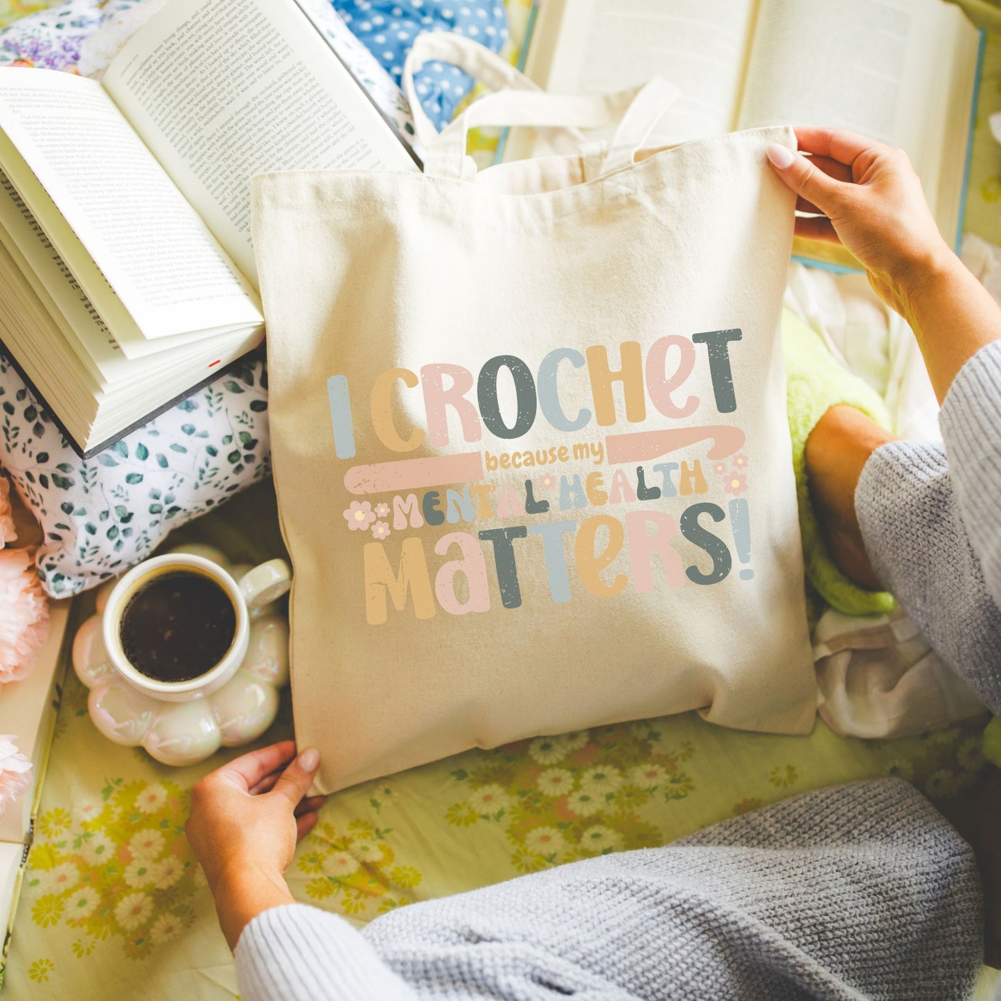 Tote bag with "I Crochet because my mental health matters" text held by a person on a floral surface with books and a mug.