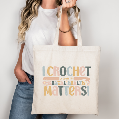 Model standing and holding a natural canvas tote with the "Crochet because my mental health matters" design.