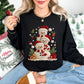 Model wearing a Gildan 18000 crewneck sweatshirt in black (Black) featuring three snowmen with faux crochet-textured white bodies, red Santa hats, and red-and-cream striped scarves, surrounded by white snowflakes, evergreen branches, wrapped gifts, a gingerbread cookie, a Christmas tree, and candy canes.