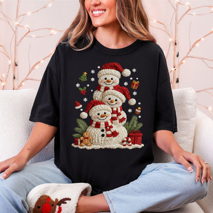 Model wearing a Comfort Colors 1717 T-Shirt in black (Black) featuring three snowmen with faux crochet-textured white bodies, red Santa hats, and red-and-cream striped scarves, surrounded by white snowflakes, evergreen branches, wrapped gifts, a gingerbread cookie, a Christmas tree, and candy canes.
