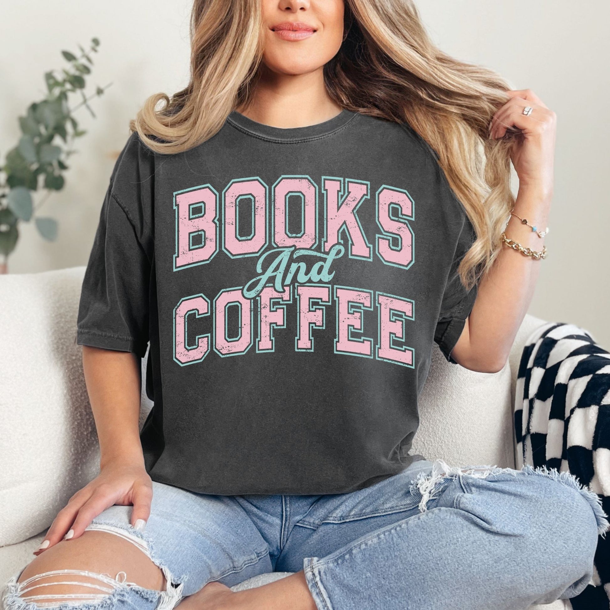 Model wearing a Comfort Colors 1717 T-shirt in a dark charcoal gray (pepper) color featuring a varsity font graphic that says, "Books and coffee" The words are light pink and outlined in pale teal green.