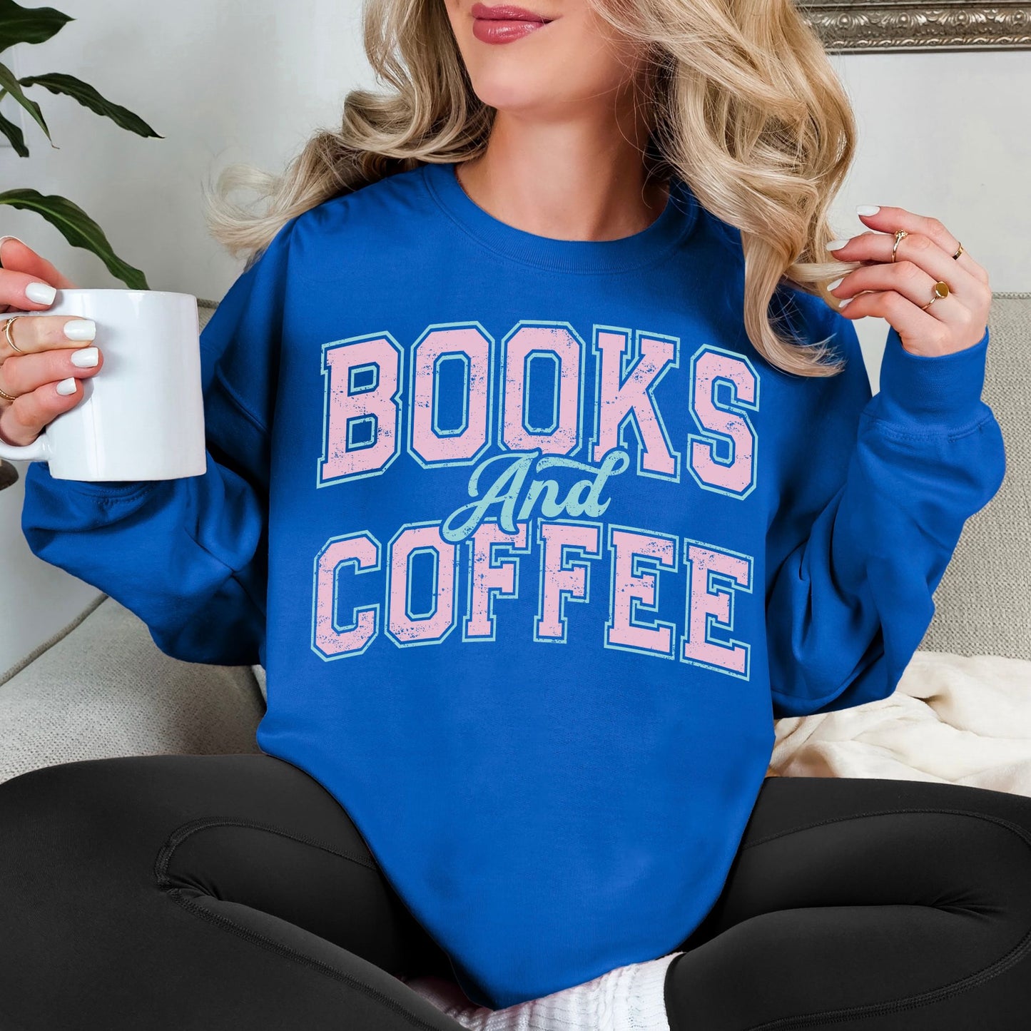 Model wearing a Gildan 18000 Sweatshirt in a bold bright blue (royal blue) featuring a varsity font graphic that says, "Books and coffee" The words are light pink and outlined in pale teal green.