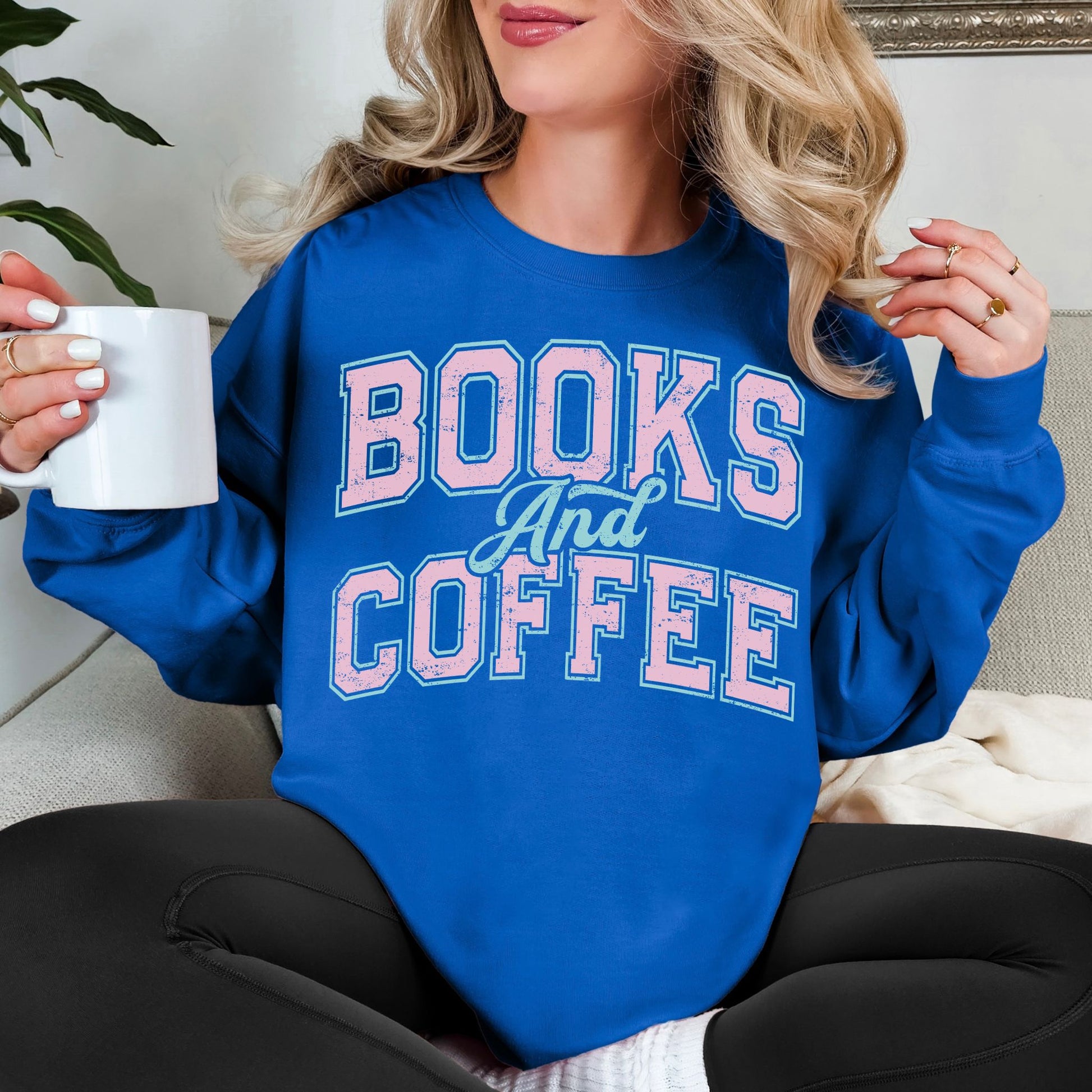 Model wearing a Gildan 18000 Sweatshirt in a bold bright blue (royal blue) featuring a varsity font graphic that says, "Books and coffee" The words are light pink and outlined in pale teal green.