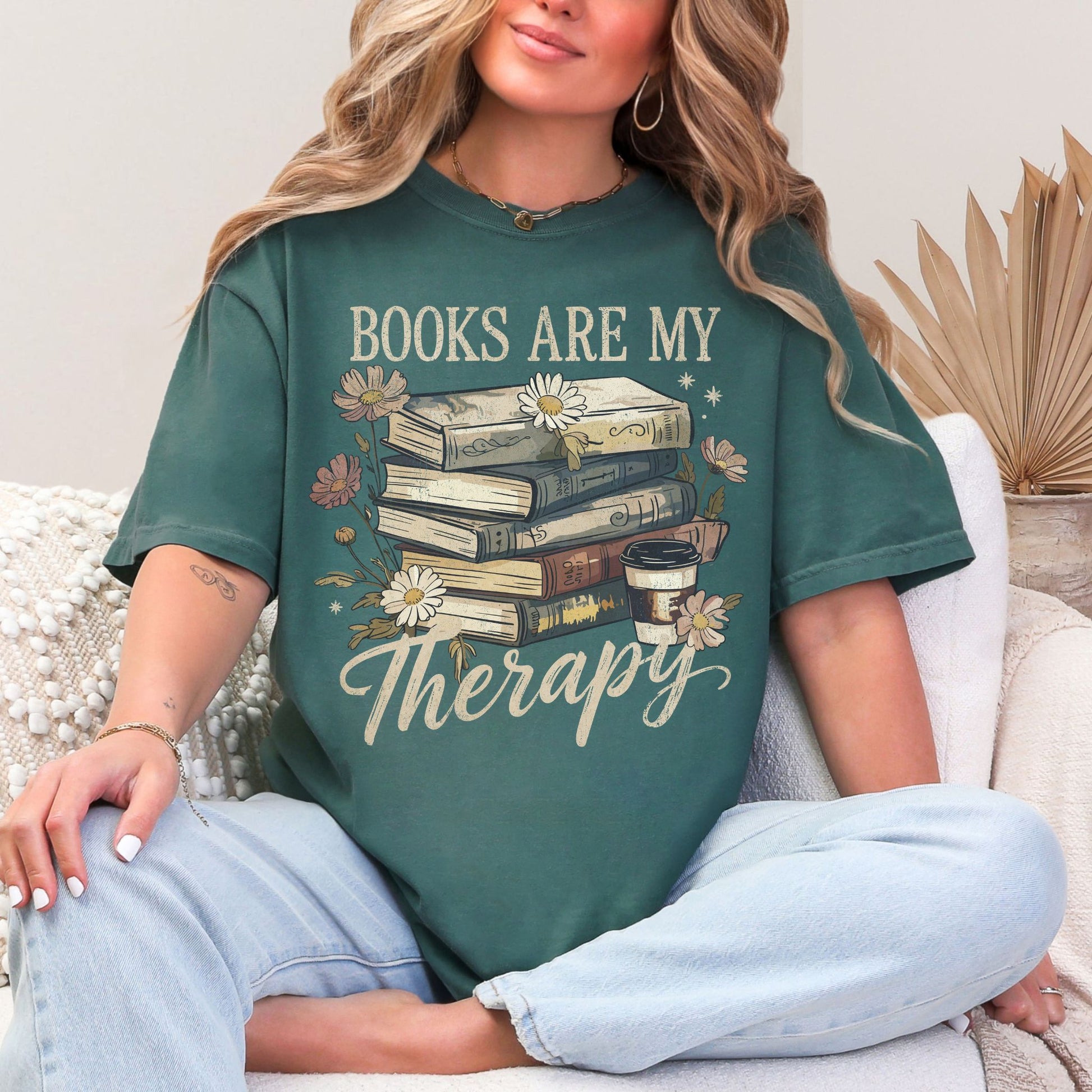 Model wearing a Comfort Colors 1717 t-shirt in a muted blue-green (blue spruce) color featuring a stacked book graphic with a to-go coffee and daisies. The phrase around the graphic is "Books are my therapy".