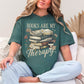 Model wearing a Comfort Colors 1717 t-shirt in a muted blue-green (blue spruce) color featuring a stacked book graphic with a to-go coffee and daisies. The phrase around the graphic is "Books are my therapy".