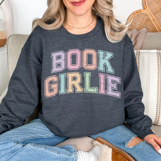 Model wearing a Gildan 18000 in a medium-dark heather gray (dark heather) color featuring distressed pastel colors of the words "Book Girlie" in a varsity letter font.