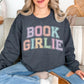 Model wearing a Gildan 18000 in a medium-dark heather gray (dark heather) color featuring distressed pastel colors of the words "Book Girlie" in a varsity letter font.