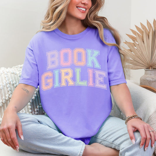 Model wearing a Comfort Colors 1717 t-shirt in a soft muted violet purple (violet) color featuring distressed pastel  colors of the words "Book Girlie" in a varsity letter font.