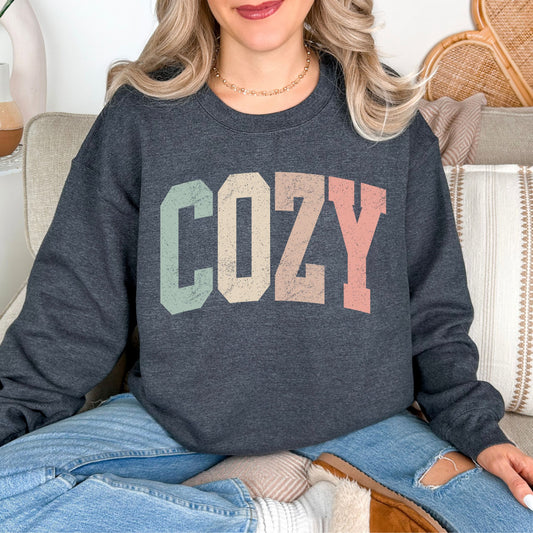 Model wearing a Gildan 18000 in medium-dark heather gray (dark heather) featuring a distressed varsity script with the word "Cozy" in pastel colors.