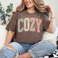 Model wearing a Comfort Colors T- Shirt in rich brown (espresso) featuring a distressed varsity script with the word "Cozy" in pastel colors. 