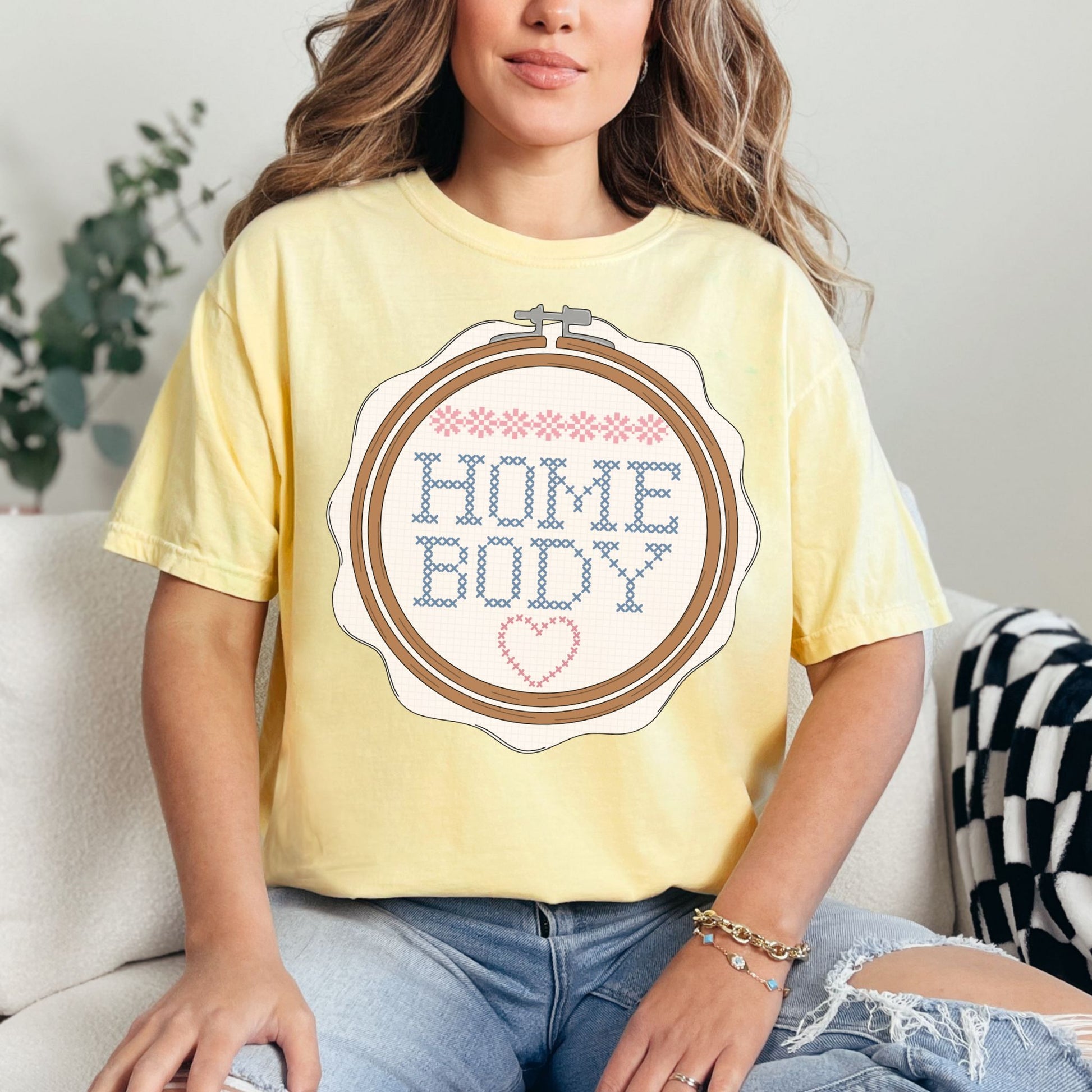 Model is wearing a Comfort Colors 1717 soft pastel yellow (banana) T-shirt featuring a faux embroidery-style design of a wooden embroidery hoop with stitched lettering that reads “HOME BODY,” accented with small pink stitch details and a heart at the bottom.
